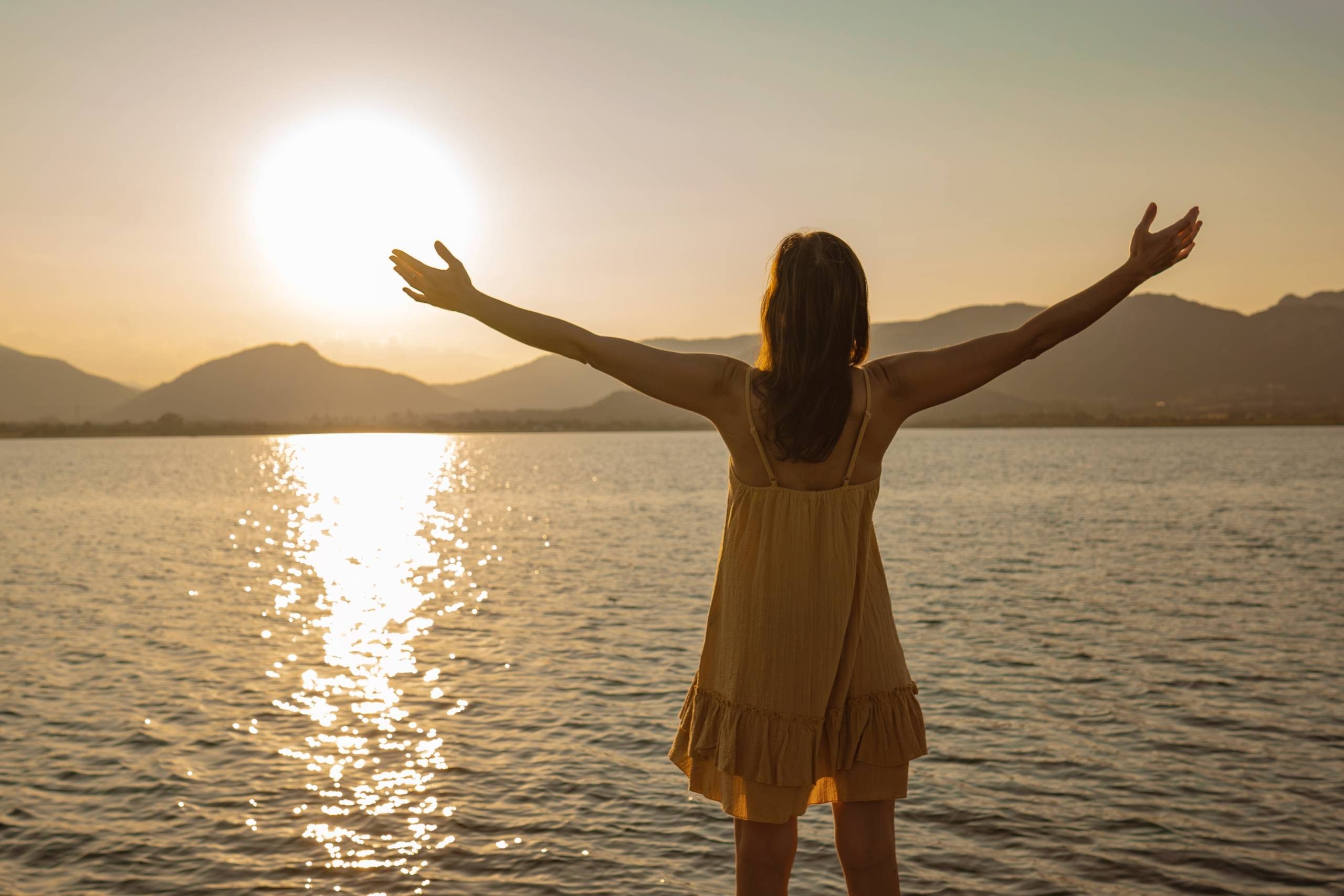 A photo of a woman in a short white dress standing on the beach facing the sun. She has her arms out facing the water. 