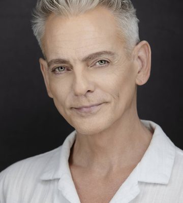 A man with platinum white hair and a white shirt stands against a black backdrop. He is smiling softly.