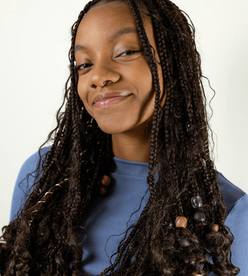 A young person with long, braided hair and beads wears a blue top and smiles slightly at the camera against a plain background.