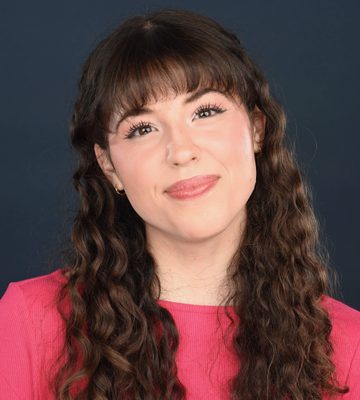 A young girl with brown curly hair wears a pink top and smiles at the camera. Her head is slightly tilted.