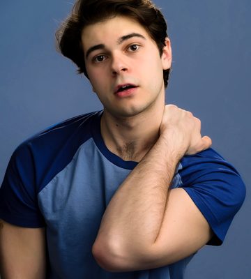 A young man in a blue ringer tee stands against a blue background. he has his hand on his shoulder and his mouth slightly agape. He has brown hair.