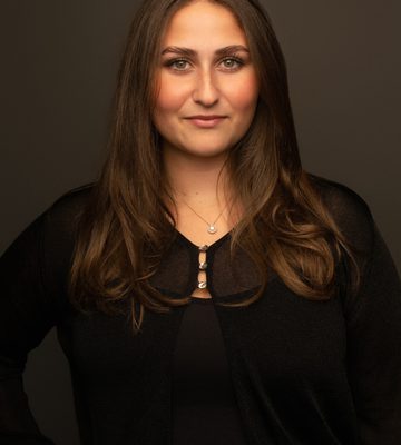 Professional headshot of a person with long brown hair, wearing a black top and black cardigan with a delicate necklace, photographed against a dark studio background.