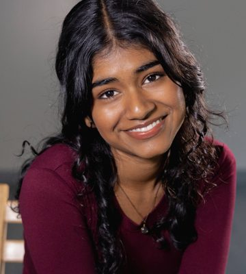 A woman with long, wavy dark hair and medium skin tone smiles at the camera. She is wearing a maroon long-sleeve shirt and a necklace, sitting on a wooden chair against a plain gray background.