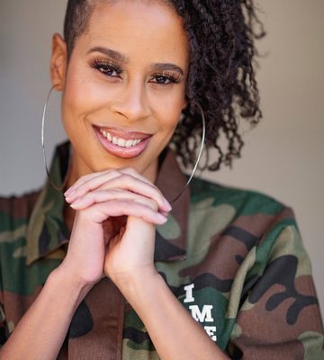 A person with hoop earrings and natural curly hair styled to one side smiles, wearing a green camouflage jacket and clasping their hands under their chin.