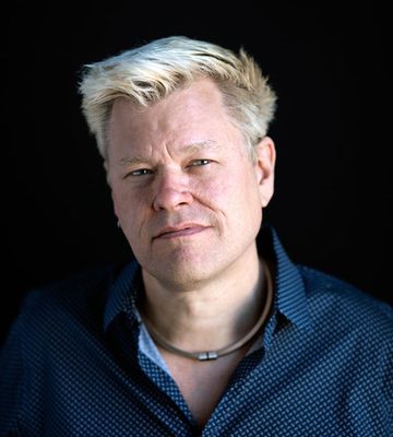 A person with short, platinum blonde hair, wearing a dark patterned shirt and a necklace, looks directly at the camera against a plain black background.