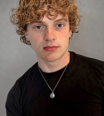 A young person with curly, light brown hair wears a black t-shirt and a silver pendant necklace, standing against a plain gray background while looking at the camera with a neutral expression.