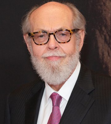 An older man with a white beard and mustache, wearing tortoiseshell glasses, a dark striped suit, white shirt, and pink patterned tie, stands in front of a dark background.