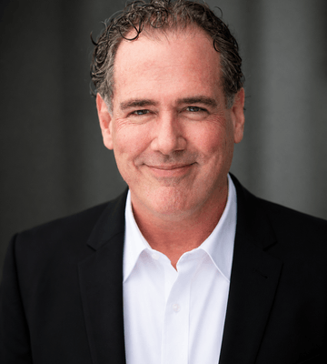 A headshot of a middle-aged white man with curly brown hair, wearing a white button-down shirt and black blazer. He is looking at the camera with a slight smile against a gray, textured background.