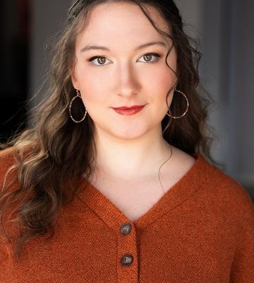 Professional headshot of a person with long wavy hair wearing hoop earrings and a rust-colored button-front sweater, photographed against a softly blurred indoor background.