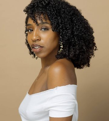Professional headshot of a person with shoulder-length curly hair wearing an off-the-shoulder white top, earrings, and a necklace, posed in profile against a soft beige studio background.