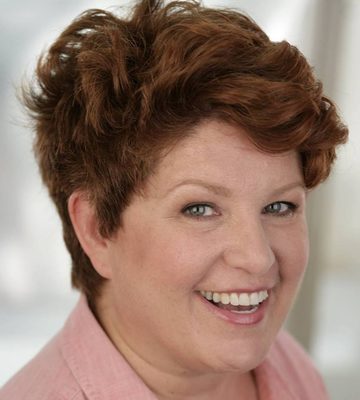 A person with short, curly reddish-brown hair smiles at the camera. They are wearing a light pink shirt. The background is softly blurred in neutral tones.