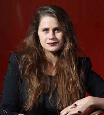 A woman with long brown hair, wearing a black blazer and black shirt, sits on a wooden chair against a solid red background, looking at the camera with a neutral expression.