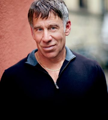 A middle-aged man with short brown hair wearing a dark zip-up shirt and gold necklace stands outdoors against a textured wall.