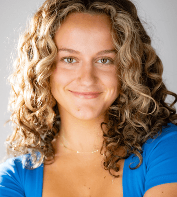 A young woman with curly hair, wearing a blue top, smiles slightly while looking at the camera. She has light skin and is shown from the shoulders up with a neutral background.