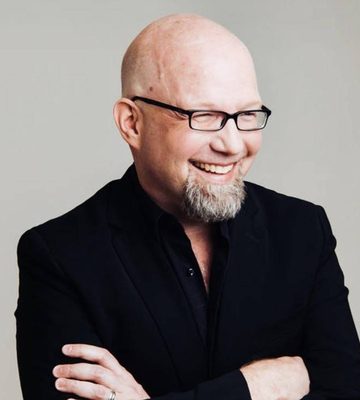 A bald man with a goatee and black-rimmed glasses is smiling while standing with his arms crossed. He is wearing a black suit jacket and a black shirt. The background is plain light gray.