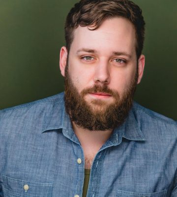 A man with a brown beard and short hair wears a blue button-up shirt over a green undershirt. He is looking directly at the camera against a plain green background.