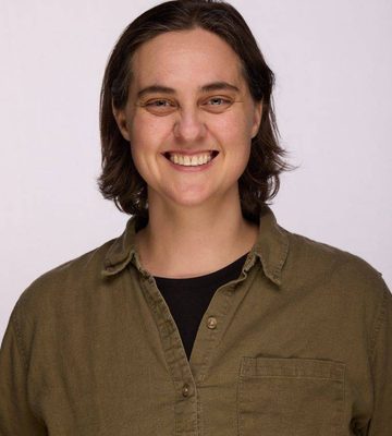 This image was uploaded directly to AltText AI for processing. A person with short brown hair is smiling at the camera. They are wearing an olive green button-up shirt over a black top. The background is plain and light-colored.