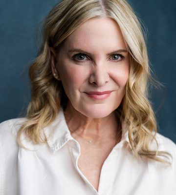 A woman with wavy blonde hair and fair skin wears a white collared shirt and smiles softly at the camera. The background is a solid, muted blue.