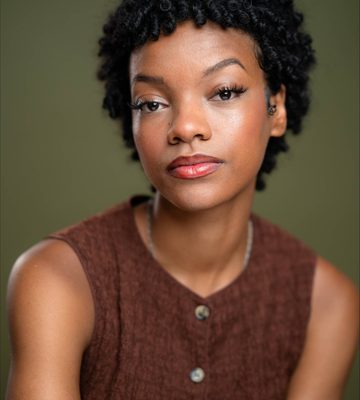 A person with short curly black hair and natural makeup wears a sleeveless brown top, looking directly at the camera with a neutral expression against an olive green background.