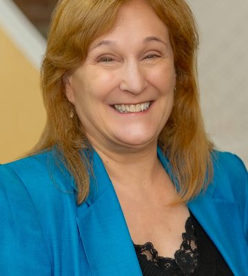 A woman with shoulder-length light brown hair is smiling. She is wearing a bright blue blazer over a black lace top. The background is softly blurred with beige and white tones.