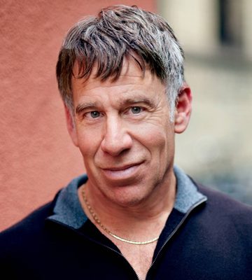 A middle-aged man with short brown hair wearing a dark zip-up shirt and gold necklace stands outdoors against a textured wall.
