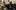 a group of people (8+) sit in chairs and stools. While you can't see them, they face an audience, appearing to be speaking towards them.