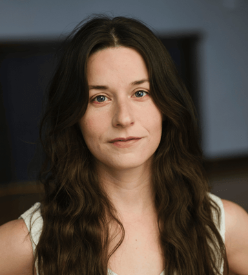 A woman with long, wavy, dark brown hair poses chest up. She wears a white tank top.