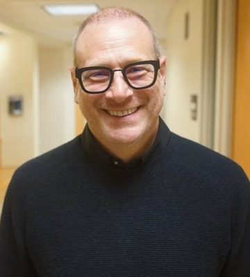 A man with glasses and a black sweater smiles at the camera in a well-lit hallway with light-colored walls.
