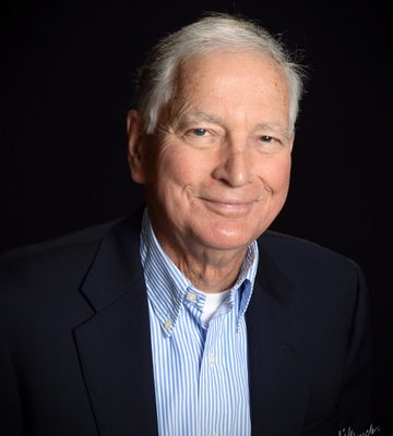 Professional headshot of a person with short white hair wearing a navy blazer over a blue-and-white striped button-down shirt. The person is seated and facing the camera against a dark studio background.