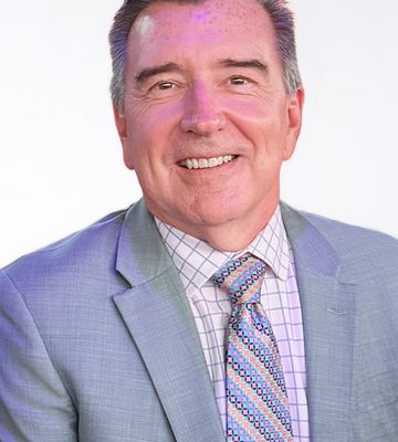 A middle-aged man in a light gray suit, patterned tie, and checkered shirt smiles at the camera against a plain white background.