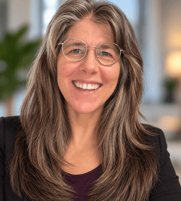 Professional headshot of a person with long, layered hair and round eyeglasses, wearing a dark blazer over a dark top. The person is centered in the frame and facing the camera. The background is softly blurred, with neutral tones and an indoor office-like setting.
