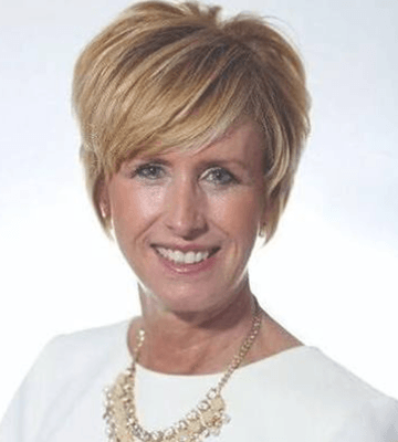 A blonde woman with shirt hair in a white shirt against a white background. She has on a gold necklace.