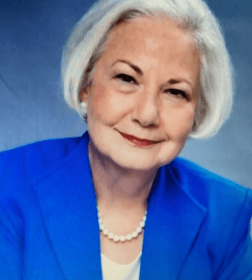 A woman in a blue jacket with a white shirt and pearl necklace against a darker blue/white ombre background.